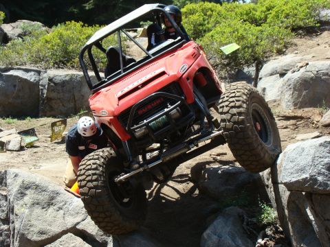 1989 Ford Bronco - A red off-road vehicle is navigating a rocky terrain with a person in a helmet assisting near the front tire. The vehicle has a roll cage a…