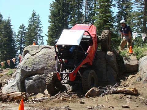 1989 Ford Bronco - An orange off-road buggy is climbing over large rocks on a dirt trail, with a person in a helmet watching. Tall pine trees are visible in t…