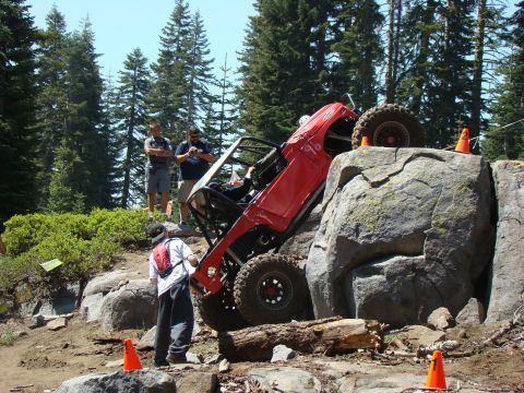 1989 Ford Bronco - A red off-road vehicle is tilted precariously on a large rock formation, with several people observing the scene. Orange cones mark the cou…