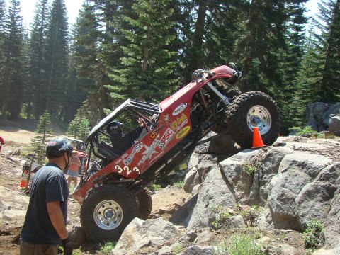 1989 Ford Bronco - A custom red off-road buggy with large tires navigates a rocky trail, with a driver wearing a helmet visible inside. The buggy is tilted si…