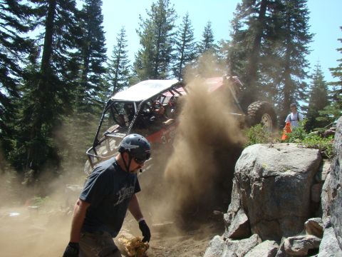 1989 Ford Bronco - A red off-road buggy with a roll cage is kicking up a large cloud of dust as it navigates a rocky, wooded terrain. A person wearing a helme…