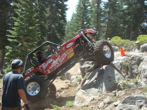 1989 Ford Bronco - A red off-road buggy with large tires is navigating a rocky terrain, with a driver wearing a helmet inside. Another person in a helmet stan…