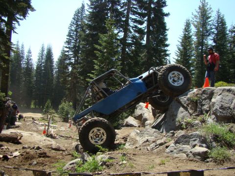 1989 Ford Bronco - A blue off-road buggy is navigating a rocky incline with its tires angled against the terrain. A man in a red shirt stands on a rock format…