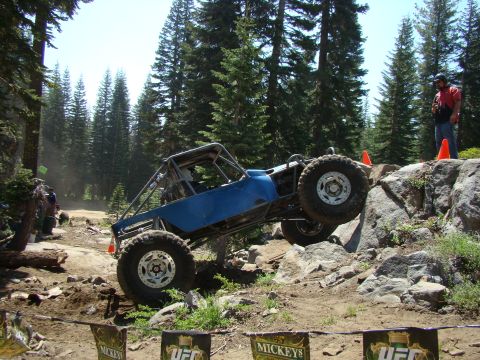 1989 Ford Bronco - A custom blue off-road buggy with large tires navigates a rocky, wooded terrain. A person in a red shirt stands on a rock formation to the…