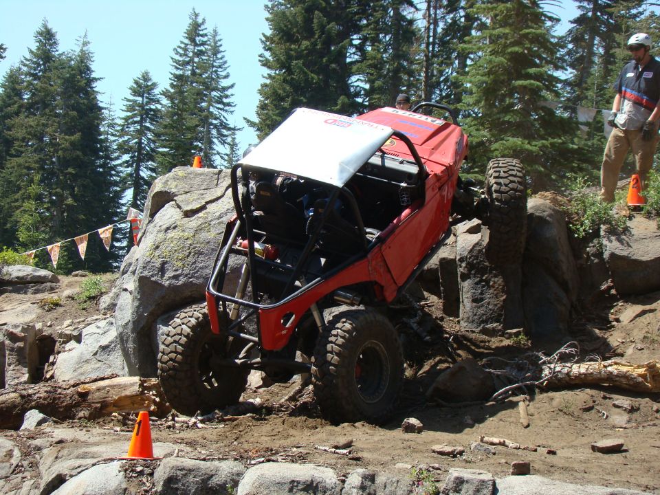 Red off-road buggy climbing a large rock obstacle in a forest.