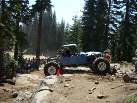 1989 Ford Bronco - A blue custom off-road buggy with large tires is navigating a rocky, wooded trail. Several spectators are visible in the background.