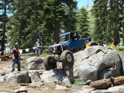 1989 Ford Bronco - A custom blue off-road buggy with a roll cage navigates a rocky obstacle course with two men standing nearby. The buggy is tilted on its si…