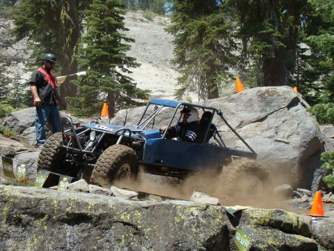1989 Ford Bronco - A blue off-road buggy navigates a rocky course, kicking up dust as it goes. A person in a helmet and vest stands nearby, observing the acti…