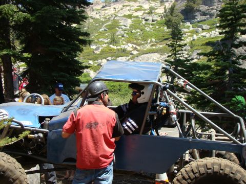 1989 Ford Bronco - Two individuals are inside a blue off-road buggy, one wearing a white helmet and sunglasses, with a mountainous, wooded background.