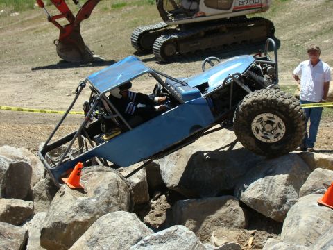 1989 Ford Bronco - A blue off-road buggy navigates a rocky obstacle course, with a spectator watching nearby.