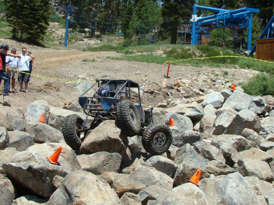 Custom off-road buggy with roll cage climbing over large rocks on a course marked with orange cones