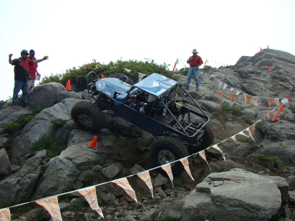 Blue off-road buggy with roll cage navigating a rocky incline, people watching
