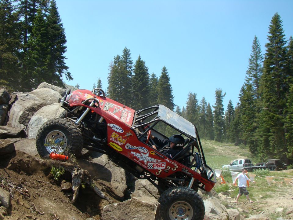Red off-road buggy with roll cage climbing a rocky incline, trees and a white pickup truck in background