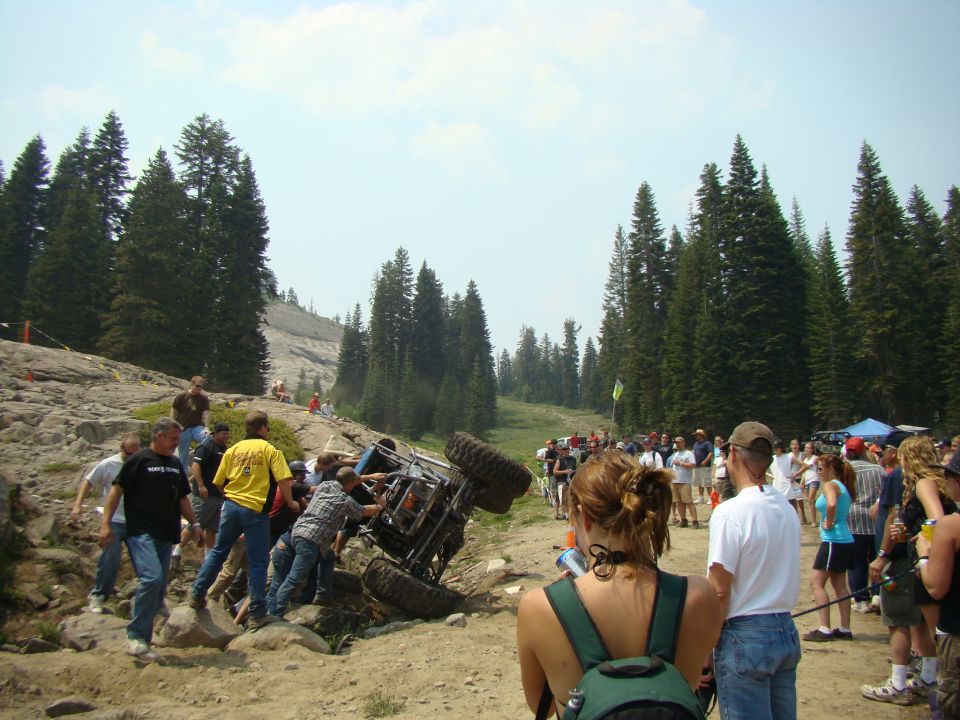 Off-road vehicle tipped on its side with people trying to help it. Crowd watches in a wooded, mountainous area