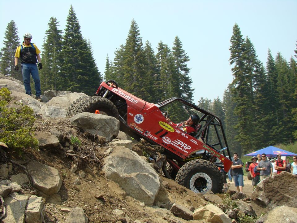 Red off-road buggy with roll cage climbing a rocky hill. Driver wears helmet. Spectators watch