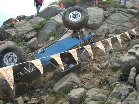 1989 Ford Bronco - An off-road buggy is tipped on its side on a steep, rocky incline, with a spare tire precariously balanced on its roof.