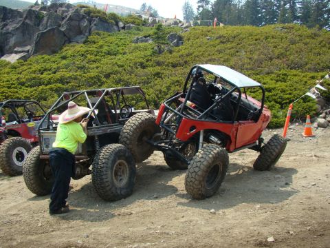 1989 Ford Bronco - Two custom off-road vehicles are parked on a dirt trail, with a person attending to one of them.