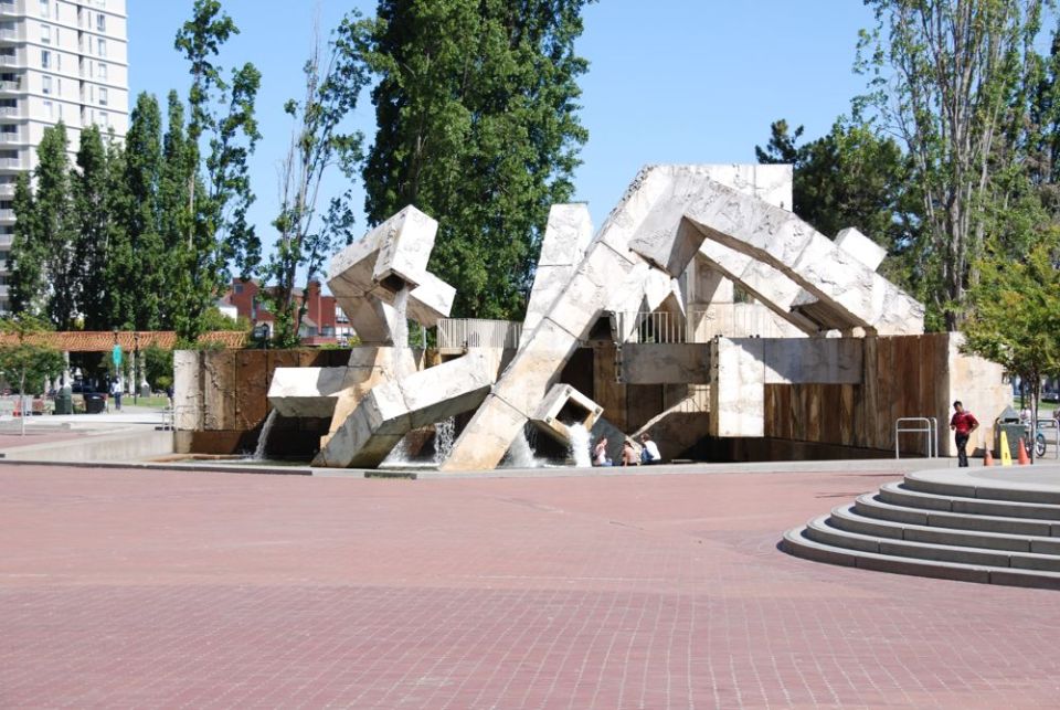 Abstract concrete sculpture with water flowing, people sitting near it, and a person standing nearby
