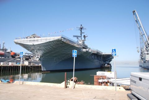 1996 Ford Bronco - A large aircraft carrier is docked in a harbor under a clear blue sky, with industrial cranes visible in the background.