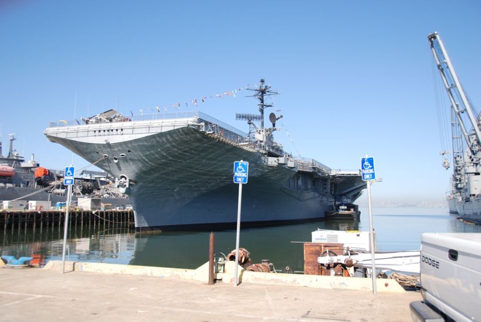 large aircraft carrier docked in harbor with cranes and a pickup truck nearby
