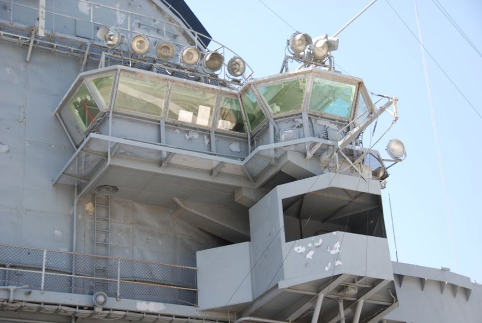 Gray ship's bridge with multiple windows and lights against a blue sky