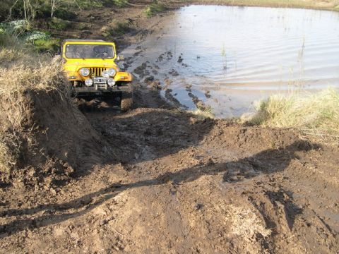 1996 Ford Explorer - A yellow off-road vehicle navigates a muddy terrain next to a calm body of water.