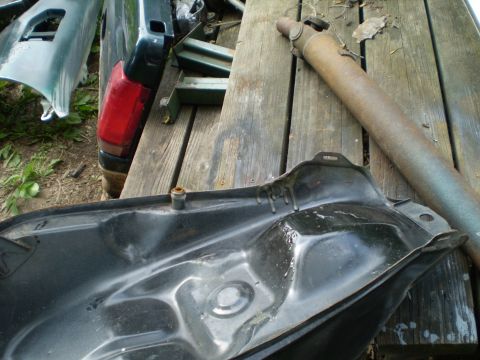1984 Honda VF700S - A black motorcycle fuel tank and a rusty driveshaft are laid out on wooden planks, with parts of a dark green vehicle visible in the backgr…