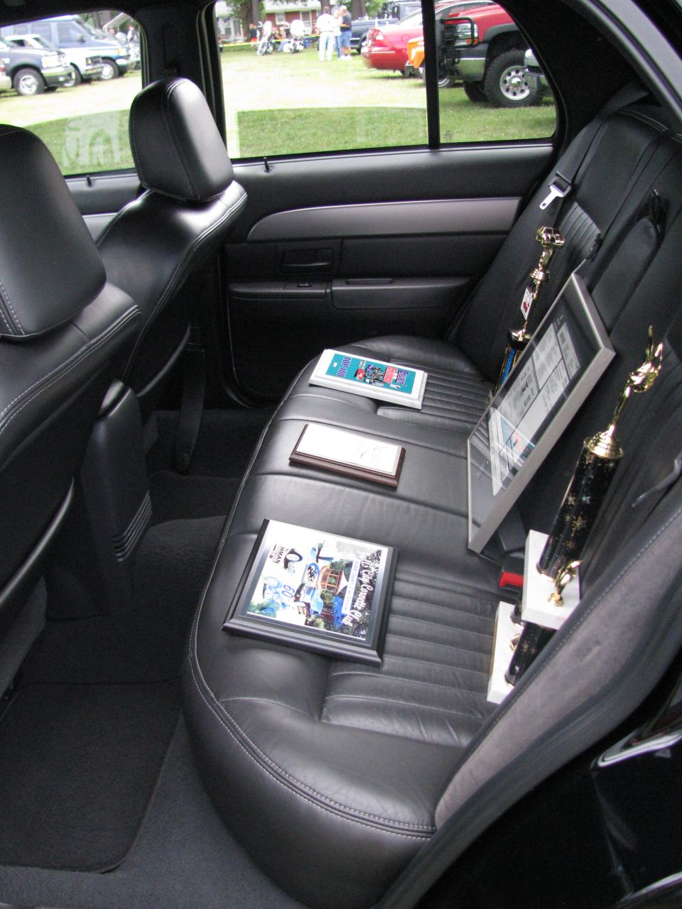 Interior view of a car's back seat with two trophies and two framed items on the seats. Other cars are visible outside the window