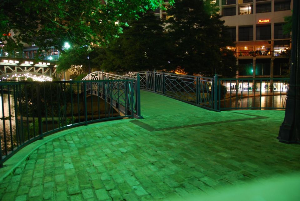 Night view of a bridge over a river, with buildings and trees in the background. The area is lit with green lights