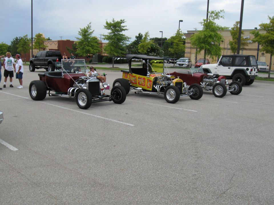 Several hot rod cars parked in a lot, including a maroon T-bucket and a yellow T-bucket with graphics. A black pickup and a white Jeep are also visible