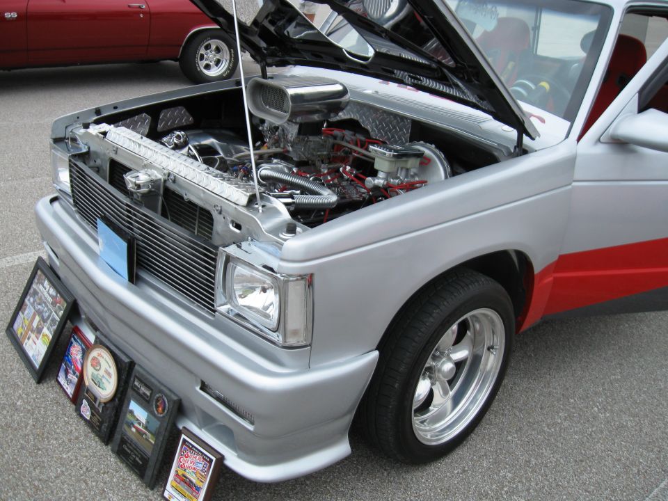 Custom pickup truck with hood open, showing engine. Awards are displayed in front of the grille. Another red car is parked behind it