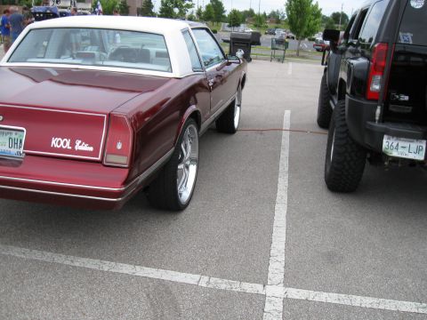 1994 Ford Bronco - A maroon coupe with a white roof and custom rims is parked next to a lifted black SUV in a parking lot.