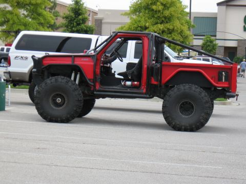 1994 Ford Bronco - A heavily modified red truck with oversized tires and a roll cage is parked in a lot.