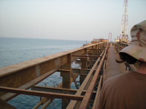 1981 Kenworth W900A - A person walks along a weathered industrial pier extending over the ocean, with a tall tower visible in the distance.
