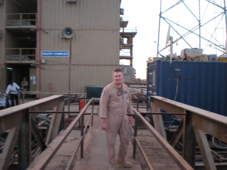 Man in tan uniform standing on a walkway with industrial buildings in the background