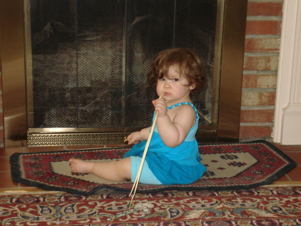 Baby in blue dress sits on rug in front of fireplace, holding sticks in mouth