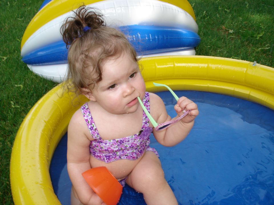 Baby in a purple floral swimsuit sits in a blue kiddie pool with a yellow inflatable ring, holding sunglasses