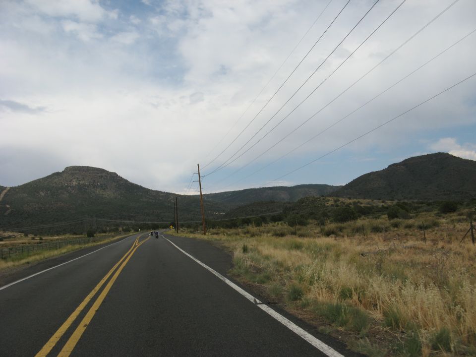 Motorcycles on a two-lane road in a rural, mountainous area under a cloudy sky