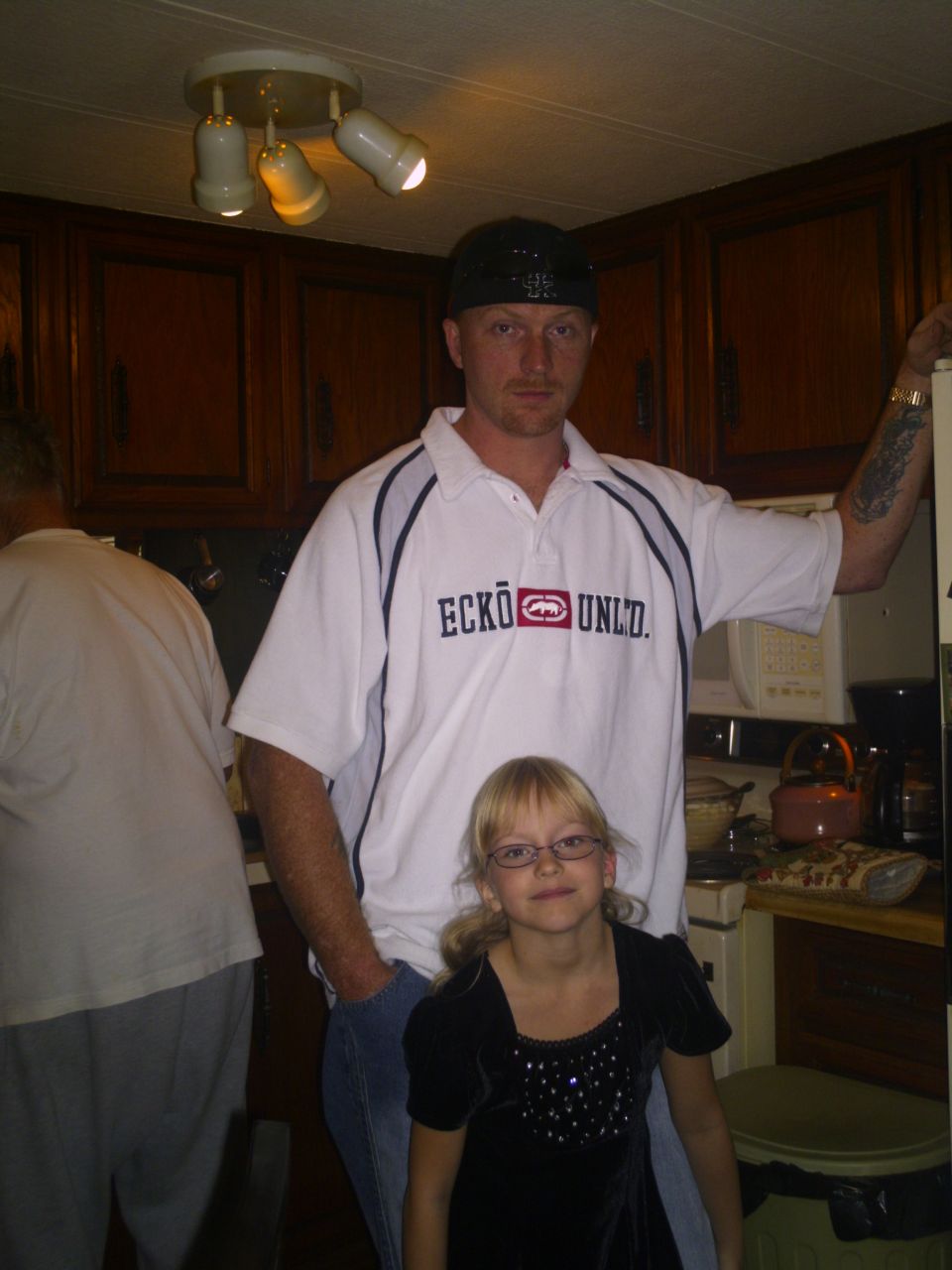 Man in white Ecko shirt and backwards cap posing with a young girl in a kitchen