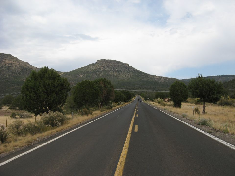 two-lane road stretches into the distance, flanked by dry grass and trees, with mountains under a cloudy sky