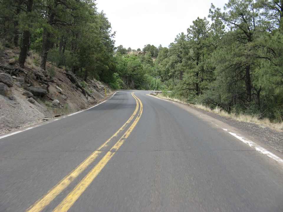 Winding mountain road with double yellow lines, bordered by trees and rocky terrain