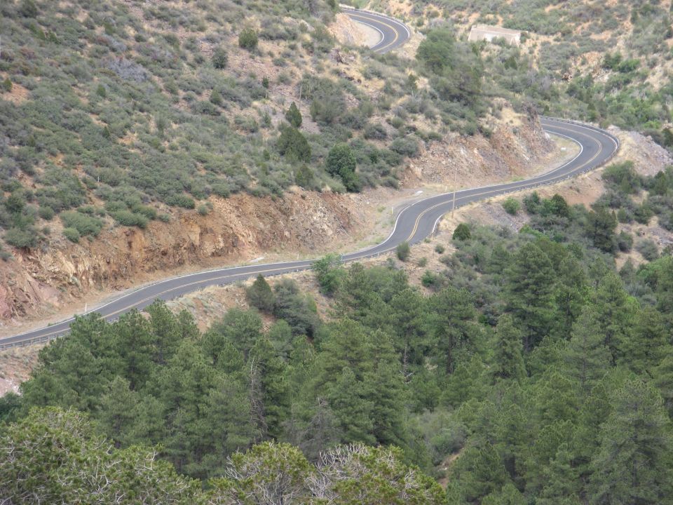Winding asphalt road through a dry, scrub-covered mountainside with pine trees in the foreground