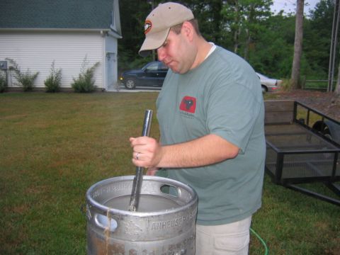1985 Ford LTD Crown Victoria - A man stirs a large metal barrel in a backyard setting, with a dark SUV and a trailer visible.