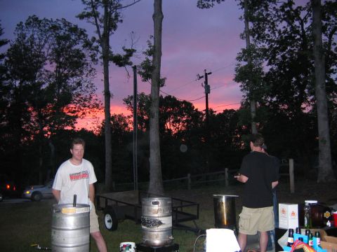 1985 Ford LTD Crown Victoria - People gather outdoors with brewing equipment and kegs as the sun sets, casting a vibrant glow across the sky.