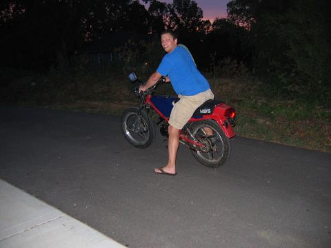 1985 Ford LTD Crown Victoria - Man on a red and blue motorcycle with MBS logo on the rear fender, riding on a paved road at dusk.