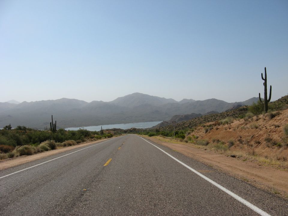 Desert highway leads toward a lake with mountains in the background. Saguaros are on the side of the road