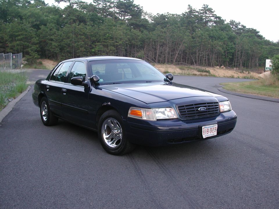 Dark blue Ford Crown Victoria parked on asphalt road, trees in background. License plate reads B1B 023