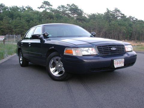 1999 Ford Crown Victoria - A dark blue sedan with chrome wheels is parked on a paved surface with trees in the background.