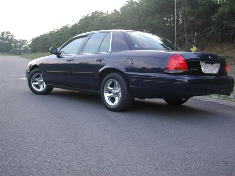 1999 Ford Crown Victoria - Rear view of a dark blue sedan on a road, with trees lining the horizon.