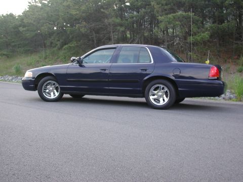 1999 Ford Crown Victoria - A dark blue sedan is parked on a road with trees in the background. The vehicle has chrome wheels and a side view is visible.